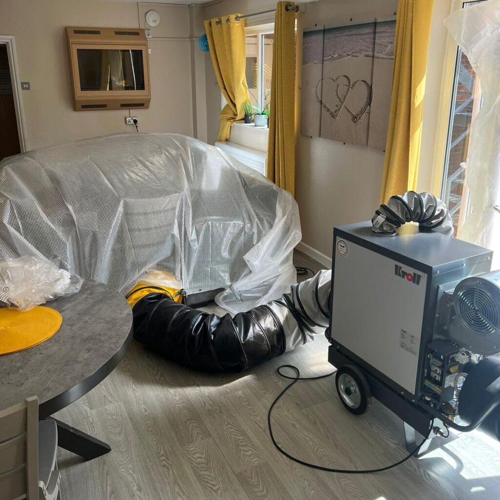 A picture of a living room with a large heat treatment machine in it. A sofa has been covered in plastic wrap to be treated.